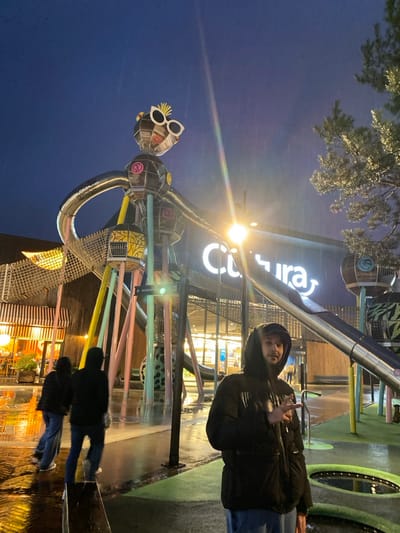 A vertical shot shows a person wearing a dark jacket and hood, flashing a peace sign. The person stands next to a colorful structure featuring a figure with oversized sunglasses. A large sign with the word "Conuray" is visible in the background. The sky is a deep blue, suggesting it is evening or nighttime. The ground appears wet, possibly from rain. Two more people are seen in the distance standing near the structure.