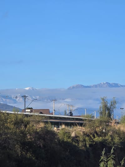 The image depicts a scene in Puente Alto, Chile, with a focus on a landscape that blends natural elements with urban infrastructure. On the right side, dense foliage and trees dominate, providing a natural backdrop. Juxtaposed against this greenery are elements of the town's infrastructure, including power lines and portions of buildings, notably the roofline of a structure with a gable roof. In the distance, a mountain range is partially obscured by clouds or fog, adding depth to the scene. The sky takes up the upper left portion of the image, presenting a clear and expansive blue.
