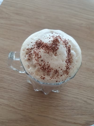 A close-up, high-angle shot shows a glass mug filled with a frothy beverage, possibly coffee or a latte, topped with a dusting of cocoa powder arranged in a pattern resembling the letter 'M'. The mug is transparent with a glass handle and a metallic rim around the top. It rests on a light brown wooden surface. The background is blurred, suggesting the image was taken indoors, potentially in a cafe or home setting. There is no visible indication of the city of Kheyrabad, Iran, or any specific geographical markers.