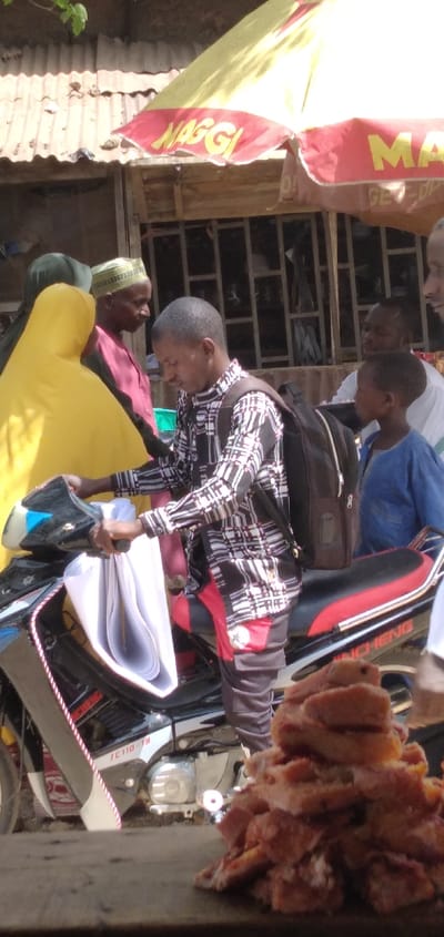 The image depicts an outdoor commercial scene in Chikaji, Nigeria, under bright daylight. A dark-skinned adult male is centered, wearing a black and white plaid long-sleeved shirt, dark grey pants with red paneling and a white star emblem on the right pocket, and a dark brown backpack. He stands alongside a black and red Jincheng JC110-1B motorcycle, holding a large, rolled bundle of white papers. To his immediate left, a female wearing a bright yellow head covering and a dark garment stands near a male with a light-colored cap and maroon shirt. To the central male's right, a young dark-skinned boy in a blue garment is visible, looking to the right. A partial view of another adult male's face is seen behind the boy. In the lower right foreground, a blurred pile of irregularly shaped, golden-brown fried food items rests on a dark surface. Overhead, a partially visible yellow and red outdoor umbrella with the "MAGGI" logo provides shade over a structure featuring a corrugated metal roof and a window with dark grilles. The ground appears unpaved.