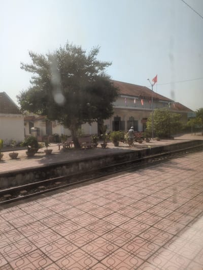 A building with a red-tiled roof is visible on the left, adorned with red and other colored flags, possibly for a local celebration. A tree stands in front of the building. A walkway paved with interlocking geometric patterned tiles runs along the side of the building. A person is near the building tending to plants. The location may be Ninh Hòa, Vietnam.