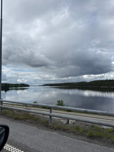 A wide shot captures a serene lake reflecting a cloudy sky, flanked by dense green forests along its shores. A road with a metal guardrail runs parallel to the water, with a portion of a car visible in the foreground. The scene is likely located in Ørje, Norway, given the characteristic landscape of lakes and forests common in the region. The sky is overcast with dramatic, dark clouds, suggesting an impending storm or recent rainfall.