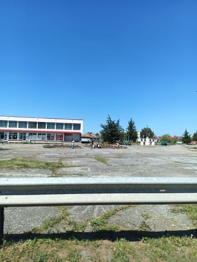 The image depicts a town square in Zimnitsa, Bulgaria. The sky is clear and blue. The foreground is a grey guardrail, with overgrown vegetation on the adjacent ground. In the middle ground, a large concrete square is visible, showing signs of weathering and sprouting vegetation. Several people are scattered across the square. To the left stands a white, two-story building with a red-trimmed roof and numerous windows. Further into the square, there is a monument surrounded by trees and two green cannons, a symbol of the village. The overall impression is of a quiet, somewhat rundown public space.