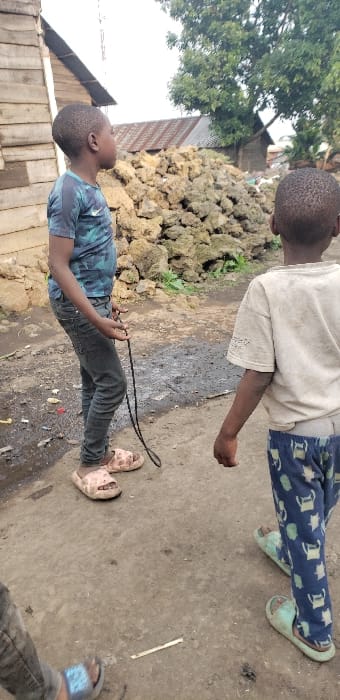 The image depicts an outdoor scene within Buzove, Democratic Republic of the Congo, on an unpaved dirt surface. Three individuals are present. In the left foreground, a young male stands facing right, his head turned upwards. He wears a blue and black camouflage-patterned short-sleeve shirt, dark grey jeans, and pink slide sandals, holding a dark looped cord in his right hand. To his right, a second young male walks away from the viewer, observed from the back, wearing a light-colored short-sleeve t-shirt with perforated sleeves, blue pants with a light-colored pattern, and light green slide sandals. The lower legs and feet of a third individual, wearing dark pants and light blue slide sandals, are visible at the bottom left. The ground exhibits compacted dirt with scattered small debris and localized dark, wet patches. In the background, from left to right, are a wooden plank structure, a substantial pile of light-colored rubble, multiple buildings with corrugated metal roofs, and a large tree with green foliage. The sky overhead appears overcast.