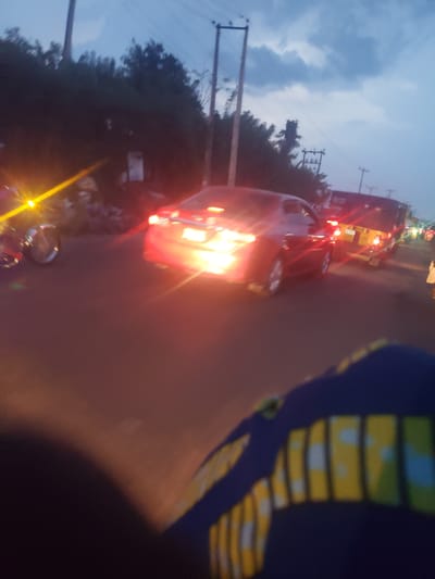 The image shows a street scene in Ida, Nigeria, likely at dusk or night. A red car is illuminated by its headlights and brake lights. A yellow van is further down the road, also with its lights on. There are streetlights and power lines visible in the background. Trees line the left side of the road. A partially visible figure wearing a blue and yellow patterned garment appears in the foreground, partially obstructing the view. Another vehicle, potentially a motorcycle, is visible near the red car. The overall scene suggests traffic and movement.