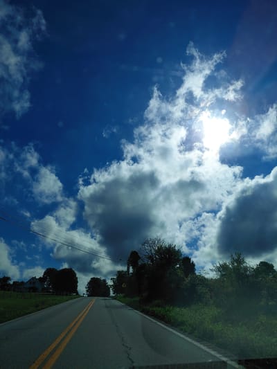 The image shows a road winding uphill under a bright, partly cloudy sky. The sun is visible, creating a lens flare effect. The road is paved and has double yellow lines in the center. On the left side of the road, there is a grassy field, a white fence, and a house with a blue roof. Trees and vegetation line the right side of the road, with some darker clouds visible in the sky. Overhead power lines are also present. The scene appears to be in a rural or suburban area, possibly in Owingsville, United States, given the style of the house and the landscape. There are no people or animals visible in the image.