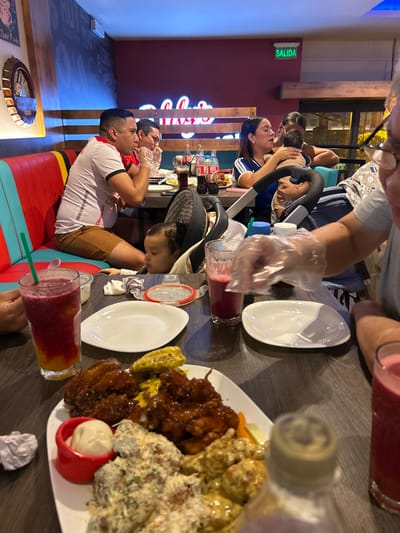 A group of people, including a baby in a stroller, are seated at a table enjoying a meal at a restaurant. They wear plastic gloves, and plates are filled with various types of chicken wings and dips. Glasses of blended drinks sit beside the plates. A sign on the wall says "Salida," and there are Coca-Cola bottles on the table. The seating area features colorful seating and decor.