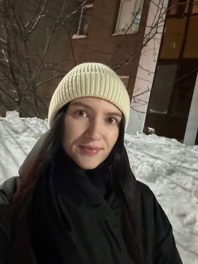 A woman with dark hair and a light-colored knit hat smiles at the camera. She's wearing a black coat, likely outdoors in the winter, as snow is visible. A building with illuminated windows is in the background, along with bare tree branches. The photo appears slightly rotated.