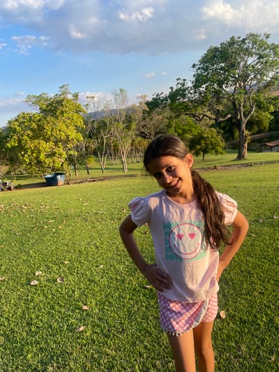 The image depicts an outdoor scene in Pirenópolis, Brazil, featuring a young female subject. The girl, appearing approximately 8-12 years old, stands centered in the foreground, facing slightly left with her body angled towards the viewer, hands on hips, and a broad smile. She has long, dark brown hair styled in a side ponytail over her right shoulder. Her attire consists of a light pink short-sleeved t-shirt with puff sleeves, emblazoned with a white smiley face with red heart-shaped eyes on a teal rectangular background, and pink shorts with white polka dots and white pom-pom trim.
The subject stands on a vibrant green grassy field, with scattered dry leaves visible on the ground. In the mid-ground, various trees are present; a large, mature tree with dense green foliage dominates the right side, while multiple smaller trees, some with sparser leaves, are arranged across the left and center. A weathered blue tub or container rests on the grass to the far left. In the background, distant hills or mountains are visible under a partly cloudy blue sky. A small structure with a brown roof is partially discernible behind the large tree on the right. The scene is illuminated by natural sunlight, characteristic of late afternoon or early morning, casting a warm glow on the foliage and grass.