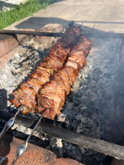 In Бучацінскі сельскі Савет, Belarus, the image shows two skewers of meat cooking over hot coals. The meat, appearing to be pork or lamb, is cut into bite-sized pieces and threaded onto metal skewers. It is visibly browned and charred in places, indicating it is nearing completion. Smoke rises from the coals, adding to the smoky aroma and visual appeal of the barbecue. The skewers rest on metal rods placed over a makeshift grill constructed with bricks. In the background, the scene transitions to a paved area and then to green grass, suggesting an outdoor setting, possibly a backyard or picnic area. The lighting suggests it is daytime, with sunlight casting shadows on the ground.