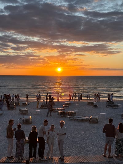 The image depicts a beach scene at sunset in Pelican Bay, United States of America. A bright, circular orange sun is low on the horizon, positioned centrally over a calm body of water, which reflects the sun's light as a golden path. The sky above features scattered dark grey and orange-toned clouds. The sandy beach is populated by approximately 50 to 100 individuals, organized in various small groups. A concentration of people stands near the water's edge, many appearing as silhouettes against the setting sun. Further inland on the sand, numerous beach lounge chairs are arranged in rows, some stacked, alongside several small white tables. In the immediate foreground, a wooden slatted platform extends, where a group of six individuals stands, engaged in conversation. The scene is illuminated by the natural light of the setting sun, casting a warm glow and long shadows.