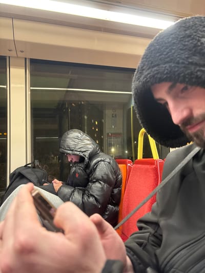 An indoor shot shows two individuals on a train or bus. One, with a grey hooded jacket, appears in the upper left corner. Another individual, wearing a glossy black puffy jacket, sits near a red seat. A large window is visible, showing the night landscape outside. The train's interior includes white walls, a yellow bar, and reflective surfaces. A hand, presumably the photographer's, holds a phone, partially obscuring the view. The photo is taken at an awkward, tilted angle.