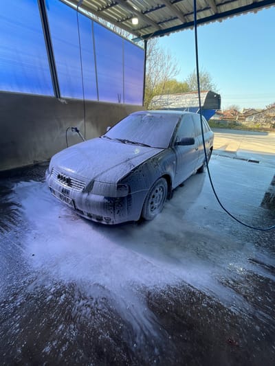 A dark car is covered in white foam at a car wash in Glojene, Bulgaria. The car is parked inside a structure with a corrugated metal roof and translucent blue panels along the sides. A black hose drapes across the top of the car. Foam and water are accumulating on the ground. The car's rear door is open. The car’s license plate reads "BH 1494 CF”. Bare trees are visible in the background.