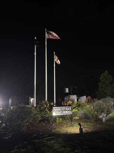 This media file depicts an outdoor scene at night, illuminated by artificial lights and a sprinkler system that appears to be in operation, casting a misty spray. The primary subjects are three flagpoles, two of which are flying flags. The topmost flag is the American flag, with its stars and stripes clearly visible. Below it, another flag is partially visible, appearing to be white with red crosses. The third flagpole is empty.
The setting is a memorial area, as indicated by a lit sign that reads "PANAMA CITY BEACH VETERAN'S MEMORIAL". Behind the sign and flags, there are structures that resemble memorial plaques or monuments, some of which are also illuminated. Lush green bushes and trees surround the area, with some flowering plants adding touches of pink to the foliage. The ground appears to be grass and dirt, wet from the sprinkler.
The time of day is clearly night, with the darkness contrasting with the illuminated elements. The atmosphere is somber and respectful, befitting a memorial site. The presence of the sprinkler suggests that the area is being maintained, possibly to water the plants or to clean the surfaces. The overall scene conveys a sense of remembrance and honor for veterans.