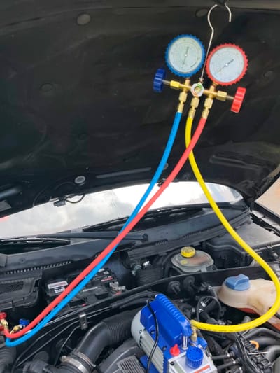 A black car hood is open, revealing the engine compartment. A set of air conditioning manifold gauges, with blue and red dials, is suspended from the hood by a metal hook. Three colored hoses – blue, yellow, and red – are connected to the gauges and extend downwards into the engine bay. A blue and silver vacuum pump is visible near the bottom right of the frame, connected to the yellow hose. The engine bay contains various components, including a car battery labeled "TUBORG TB562-051", a yellow-capped fluid reservoir, and a blue-capped fluid reservoir. The overall scene suggests a car maintenance activity, likely related to air conditioning servicing, taking place outdoors. The lighting suggests daytime. Visible text on the hoses indicates "CHARGING HOSE" and pressure ratings.