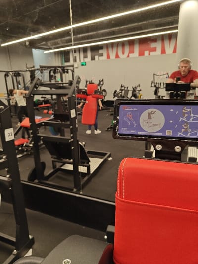 The image depicts the interior of a fitness center, specifically a gym, located in Nahariya, Israel. The perspective is from an individual seated on red upholstered gym equipment, possibly a leg press or shoulder press machine, facing a large reflective surface that constitutes a wall.

In the immediate foreground, to the right, is a red padded seat or backrest with white stitching. Directly ahead, attached to the gym equipment, is a black screen or instruction panel displaying exercise diagrams and text. Visible text includes "CalfLeader ES4" and "LB-2003 BOTH WAY SQUAT," along with illustrations demonstrating leg exercises.

The main body of the image shows the reflection of the gym's interior. The floor is covered in black rubber matting. Various pieces of gym equipment are visible, including weight machines, benches, and stationary bicycles, arranged throughout the space.

Several individuals are present in the reflection:
*   To the left, a person wearing a white tank top and dark pants is partially visible, appearing to be engaged with a weight machine.
*   In the center-midground, another individual in dark attire is positioned at a machine with prominent red padding, possibly a squat machine, facing away from the camera.
*   To the right, a male individual with short hair, wearing a red shirt and over-ear headphones, is seated on a stationary bicycle, looking down at a mobile phone.

The background wall, as seen in reflection, is light-colored. A large red graphic with inverted text, reading "HT EVOL EW" (likely "WE LOVE THE" or similar), is visible on this wall. The ceiling features multiple long, linear fluorescent light fixtures. A black structural beam runs horizontally across the bottom of the reflective surface. A small white square with the number "22" is affixed to a black metal frame in the reflected view, near the left.