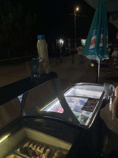The image depicts an outdoor commercial scene at night in Kostievo, Bulgaria. In the foreground, an illuminated ice cream display freezer with a partially open curved glass lid reveals packaged ice cream products and a branded "NUII" menu. To the left, a second, lower freezer section contains scoopable ice cream. On a dark counter surface to the left, a yellow plastic bottle with a blue cap stands upright next to a clear container holding multiple colorful, twisted plastic utensils. To the immediate right of the display freezer, a beige, textured object, possibly a woven hat, rests on a white, textured surface, adjacent to a crumpled clear plastic bag.
In the midground, several individuals are visible walking on a paved street; one person is seen from behind wearing a white "The North Face" t-shirt. A large, teal market umbrella, folded and displaying a white "es" logo with a snowflake graphic, occupies the right side of the midground. The background is dark, revealing dark trees to the left, distant structures, and multiple artificial light sources, including a prominent street lamp emitting a bright glow. The overall scene indicates a late-evening vendor operation in a populated area.