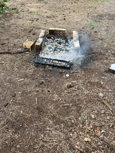 This image depicts an outdoor scene in Rudka, Ukraine. The central focus is a makeshift fire pit constructed with bricks, containing a substantial amount of ash and a partially burned log. Wisps of smoke are visible rising from the pit, indicating recent or ongoing combustion. The ground surrounding the fire pit is covered in soil, dry pine needles, twigs, and small pine cones, suggesting a natural, possibly forested or park-like environment. Two hollow bricks, appearing weathered and slightly discolored, are placed to the left of the fire pit. One brick is upright, and the other is laid on its side. The overall impression is of a rustic, possibly designated, outdoor cooking or gathering area. No individuals are present in the frame. The lighting suggests an overcast or shaded day.