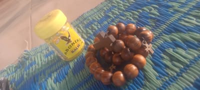 The image shows a jar of Aboniki balm and a wooden rosary resting on a blue and green woven mat. The background suggests an outdoor setting in Bende, Nigeria, indicated by the brand of the balm commonly found in Nigeria. No people are visible.
