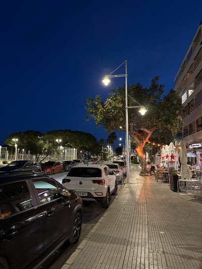 The image shows a street scene in Altea, Spain at night. A row of parked cars lines the right side of the street, closest to the viewer. In front of the cars, a pedestrian walkway is lined with lampposts and mature trees. Across the walkway, tables and chairs, covered by white parasols, suggest an outdoor restaurant or café. The sky is a deep blue, and the lighting creates a warm glow in the restaurant area. The building adjacent to the restaurant has multiple floors with lights visible through the windows.
