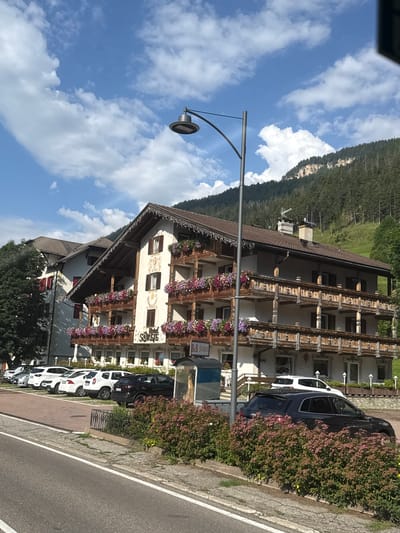 The image captures a view of the Hotel SORETE in Pozza di Fassa, Italy, under a bright blue sky with scattered white clouds. The hotel, a multi-story building with traditional alpine architecture, features wooden balconies adorned with abundant flower boxes filled with colorful blooms. The facade of the hotel is white with decorative elements, and signage indicates it is also a B&B. In front of the hotel, a row of parked cars, including white sedans and SUVs, and a black SUV are visible. A street lamp stands prominently in the foreground. The scene is bordered by a paved road and a sidewalk with a low metal fence, behind which lush green bushes and trees are planted. The background reveals a densely forested mountainside, characteristic of the Trentino-Alto Adige region.