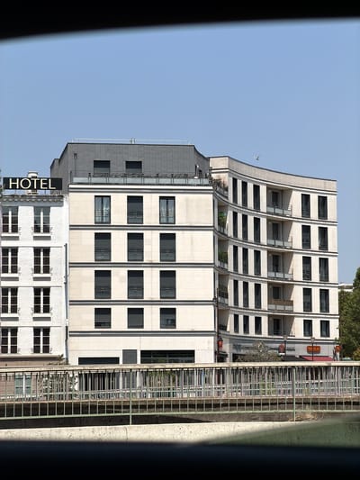 This media file captures an outdoor scene featuring a modern, multi-story hotel building. The building's facade is predominantly white with a repeating pattern of dark-framed windows and balconies. A large, dark "HOTEL" sign is prominently displayed on the lower section of the building. To the right of the hotel, a metal fence runs along the side of the structure, separating it from what appears to be a walkway or elevated path. Behind the fence, trees and some greenery are visible, suggesting an urban environment with some natural elements. The sky is clear and blue, indicating daytime and fair weather. The perspective suggests the photograph was taken from a slightly elevated position looking across at the hotel. There are no people visible in the frame. The time of day appears to be morning or afternoon, given the lighting and clear sky. A sign for "NICOLAS" is visible on an adjacent building or section of the hotel.