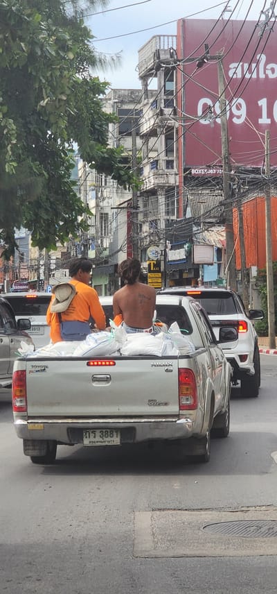 A horizontal, slightly skewed image shows a street scene in Nathon, Thailand, with a focus on a silver pickup truck overloaded with white sacks in its bed. Two individuals are partially visible in the truck bed. The person in the foreground, wearing an orange shirt and a light-colored hat, is facing away from the camera. Behind them, a shirtless person with tattoos on their back is also seated in the truck bed.
The truck is on a paved road, with other vehicles visible behind it, including a white SUV and part of another car. The background is a busy urban environment with buildings, overhead electrical wires, and greenery. The buildings appear to be a mix of old and new structures, some with weathered facades. A large red sign with Thai text and white numbers "0909 10" is prominent on the left side of the image. Other visible text includes "EXCHANGE", "HP", "9913", "DEBON", "BOXES083 70", and a license plate that reads "กร 3881". The scene suggests a daytime setting, possibly during daylight hours, with a slightly overcast or hazy sky. The overall impression is one of a bustling, working street in a tropical location.