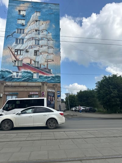 The image shows a multi-story brick building with a large mural of a sailing ship on its facade, located in Orsk, Russia. The mural depicts a red-hulled ship with white sails against a blue, wave-filled ocean and sky. Below the mural, on the ground floor, are storefronts including "WILDBERRIES" and "OZON," as well as an advertisement for "DAVRON" men's haircuts. A white passenger van and a white sedan are parked on the street in front of the building. A person in a patterned shirt and blue jeans is standing in a parking area to the right of the building, near several other parked cars and a densely wooded area. Power lines are visible against the sky. The street in the foreground has tram tracks.