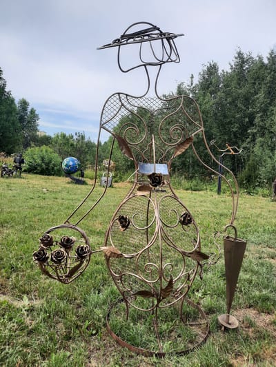 A life-sized metal sculpture of a woman stands in a grassy, outdoor area. The sculpture is made of wire and mesh, with decorative metal leaves and roses adorning its body. The woman is wearing a wide-brimmed hat and holding a basket of roses in her left arm and a cone-shaped object, possibly an umbrella stand, in her right hand. In the background, there are trees, a globe on a stand, a motorcycle with a robot figure, and a large padlock sculpture. The sky is overcast. The sculpture is located in an open field that appears to be part of a park or sculpture garden.