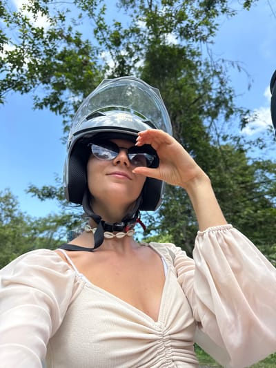 A woman wearing a motorcycle helmet and sunglasses poses for a selfie outdoors. She has a seashell necklace around her neck and is wearing a light-colored ruched top with sheer balloon sleeves. Her hand is raised to adjust her sunglasses. The background shows a blue sky with green trees. The image was taken in Ham Thuan Bac, Vietnam.
