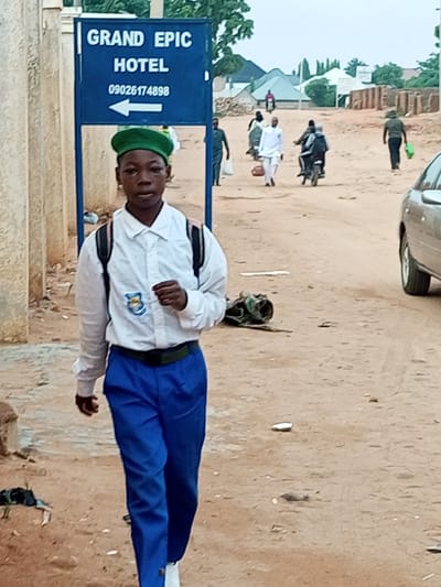 The image depicts a young person wearing a white collared shirt, blue trousers, and a green cap, walking towards the viewer on a dirt road in Wuro Dole, Nigeria. The individual carries a dark backpack. Behind them, a blue sign reads "GRAND EPIC HOTEL" with the phone number "09026174898" and an arrow pointing left. In the background, other individuals are visible, including some on motorcycles and walking. Buildings with corrugated roofs and brick structures are present, along with some vegetation. A silver car is parked on the right side of the road. The environment appears to be a dusty, rural or semi-urban area.