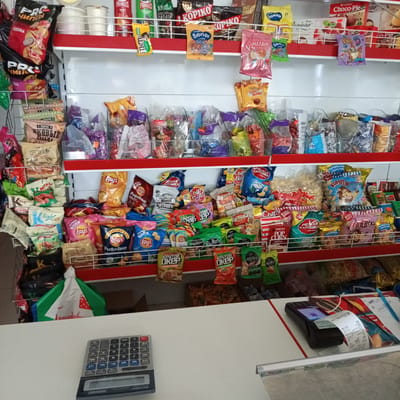The image shows a close-up view of a shelf in a small convenience store, likely located in Gorkaya-Balka, Russia. The shelf is filled with a variety of snack products, including chips, crackers, and candies, predominantly in colorful packaging. Some of the brands visible include Lay's, Pringles, and Kopiko. The products are arranged on multiple tiers, with some items hanging from hooks. In the foreground, on the counter, a calculator and a payment terminal are visible, suggesting that transactions are being processed. The overall environment appears to be a retail setting, with bright lighting and a display of consumer goods. There are no people visible in the image.