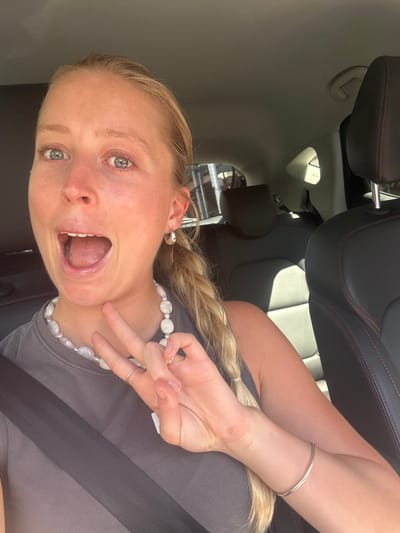 The image displays a fair-skinned female with blonde hair, styled in a braid over her left shoulder, seated within a vehicle. Her blue eyes are wide, and her mouth is open, revealing teeth. She is wearing a dark grey sleeveless top and a black seatbelt diagonally across her chest. Accessories include a necklace composed of large, irregular white pearls, small silver hoop earrings, a silver ring on her right ring finger, and a silver bracelet on her left wrist. Her right hand is raised, with the index and middle fingers extended in a "peace" sign. The vehicle's interior features black leather seats with red stitching visible on the front passenger headrest and seat edge. Bright daylight illuminates the scene, and reflections of urban structures are discernible through the rear window. The scene is located in the city of Wollongong, Australia.