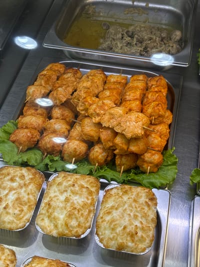 The image shows a close-up, eye-level view of food items displayed in what appears to be a refrigerated display case, likely in a food establishment in Vladikavkaz, Russia. In the foreground, three rectangular aluminum foil pans are filled with a creamy, baked dish topped with a golden-brown, slightly crispy crust, possibly a gratin or shepherd's pie. To the left and slightly behind these pans, a tray is filled with several skewers of what appear to be chicken kebabs. The chicken pieces are uniformly cut, coated in a reddish spice blend, and skewered on thin wooden sticks. These kebabs are arranged on a bed of fresh green lettuce leaves. In the background, another large metal pan contains a stew-like dish with chunks of meat and a greenish sauce. The overall impression is of a prepared food counter offering a variety of dishes. The lighting in the scene is artificial and highlights the textures and colors of the food.