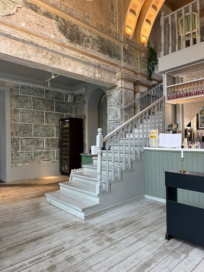The image shows an interior space with a mix of architectural styles. There's a grand, old-style wall with ornate details and weathered texture, contrasted by a modern-looking staircase with white balusters. The staircase leads to an area with light-colored plank flooring and what appears to be a menu display, near a kitchen or service area. Under the staircase is a wine display. The overall scene suggests a blend of historical and contemporary design elements. It's not possible to determine from the image if this interior is located in Перовское сельское поселение, Ukraine.