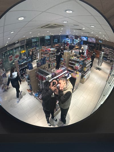 The image depicts the interior of a convenience store, specifically a gas station mini-market, located in Gorno Aleksandrovo, Bulgaria. The scene is captured through a convex security mirror, resulting in a wide-angle, distorted view within a circular frame.
In the immediate foreground, reflected in the mirror, two females are positioned closely together. The individual on the left, wearing a dark puffer jacket and dark pants with black and white Nike slides, holds a dark-colored smartphone vertically, appearing to take a selfie or photograph. The individual on the right, wearing a light-colored puffer vest over a dark top and dark pants with light-colored slides, has her left hand raised to her lower face and is looking towards the phone.
The store's interior features a white tiled floor and a suspended ceiling with multiple recessed lights and an air conditioning unit. Merchandise is displayed on numerous aisles and shelves, stocked with items such as packaged snacks, beverages, and other consumer goods.
Multiple individuals are visible within the store. On the left side, a person in a black and white tracksuit walks away from the mirror's perspective, near a display with a "Red Bull" logo. Towards the center, several people are gathered around a counter area, one person in a light-colored top standing behind it, possibly a store employee. Additional customers or staff are present further back in the store near refrigeration units. Large glass windows or doors are visible at the far end of the establishment, revealing an indistinct exterior view, possibly at night.