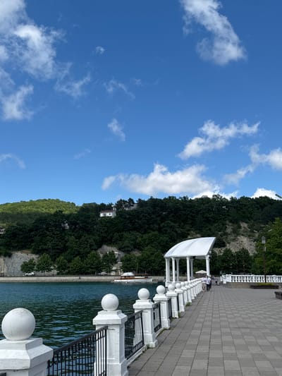 This is a sunny day photo taken at the promenade in Abrau-Dyurso, Russia, overlooking the Abrau Reservoir. In the foreground, a white railing with spherical finials lines a paved walkway. Beyond the railing, the calm, blue water of the reservoir reflects the sky. Across the reservoir, a steep, tree-covered hill rises, dotted with white houses. On the promenade, two individuals walk towards a white gazebo structure supported by columns. The sky is a vibrant blue with scattered white clouds.