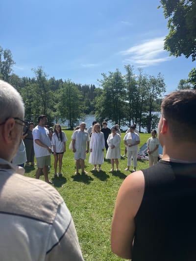 A group of people are gathered outdoors on a grassy area near a body of water, likely for a ceremony or event. The weather is sunny with a few clouds in the sky. The environment includes trees and a lake or large pond in the background. Most of the people in the central group are wearing white clothing, suggesting a shared occasion. Some individuals are speaking or listening intently, and one person is holding a microphone. The scene appears to be taking place on a warm, pleasant day. The precise location in Espoo, Finland, is not discernible from the image.