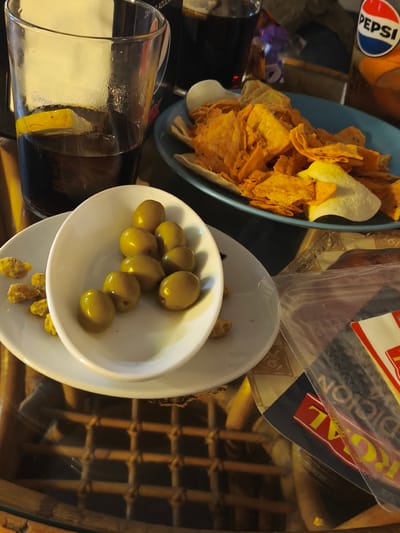 The image shows a table with a casual spread of food and drinks. A glass containing a dark liquid and a lemon slice sits next to a blue plate filled with tortilla chips. In the foreground, there's a small white bowl with green olives, placed on a larger white plate that also holds some yellow-brownish small pieces. A bottle of Pepsi and another glass with a dark beverage are visible in the background. A partially visible package with the word "ROYAL" is also on the table. The table itself has a woven, possibly rattan or wicker, base, visible through a glass top. Although the location isn't explicitly shown, the presence of Spanish food and packaging suggests the scene is likely in Spain, possibly in Moaña.