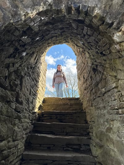 The image captures a single female subject standing at the top of a stone staircase, framed by the illuminated exit of a darkened, arched stone passage. This structure is consistent with historical ruins such as those found at Blankenstein Castle (Burg Blankenstein) in Hattingen, Germany.

The female, possessing medium-length dark hair, is dressed in a light brown puffer-style jacket, light blue jeans, and light-colored shoes. She is positioned facing the viewer, with a slight smile, hands resting at her sides.

The immediate environment consists of a rough-hewn stone tunnel or archway and a series of uneven, irregular stone steps ascending towards the exterior opening. The interior of the passage and the lower steps are largely in shadow, while the upper steps and the female figure are brightly illuminated by natural light entering from the archway.

The background visible through the archway consists of a blue sky with scattered white clouds. The bare branches of deciduous trees are visible against the sky, suggesting a leafless season. No other individuals or significant objects are present in the scene.