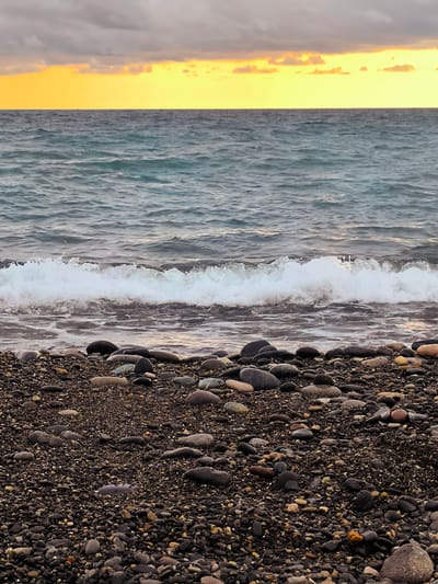The image depicts a rocky beach scene with the Black Sea in Zemo Kvariati, Georgia. The foreground is dominated by a dense scattering of dark, smooth pebbles and stones, with some larger, rounded rocks interspersed. A gentle wave is breaking on the shore, its white foam contrasting with the darker stones. The water is a deep, muted blue-green, showing ripples from the movement. In the background, a bright, warm yellow band of sky is visible just above the horizon, tinged with orange and gray clouds overhead, suggesting either sunrise or sunset. There are no people or man-made objects visible in the frame.