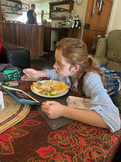 The media file shows a young person with long, reddish-brown hair, wearing a blue and white striped shirt, leaning over a table. They are eating from a plate with a fork, and their meal appears to include toast, avocado, and possibly cheese. A tablet is propped up in front of them, displaying an image on its screen.

The scene is indoors, likely a dining or living area. A patterned tablecloth covers the table. To the left, a couch with a blue and white patterned throw is visible. In the background, a wooden staircase and a kitchen counter with appliances can be seen, suggesting a home environment. The lighting appears to be natural, possibly from a window out of frame, indicating it's daytime. A person wearing a black tank top is partially visible in the background, standing near the kitchen area. The overall mood seems casual and relaxed, with the person focused on their meal and the tablet.