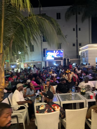 The image depicts a lively outdoor evening gathering at a venue in Oba, Nigeria. A large screen displays a vibrant image, likely a performance or broadcast, drawing the attention of many attendees. The scene is populated with numerous individuals seated at tables, engaged in conversation and enjoying the atmosphere. Palm trees are visible, suggesting a tropical or sub-tropical environment. The lighting indicates it is nighttime, with artificial lights illuminating the gathering. People are dressed in casual to semi-formal attire. The tables are set with drinks and bottles, indicating a social event with refreshments. The overall impression is one of a popular social hub with a festive ambiance.