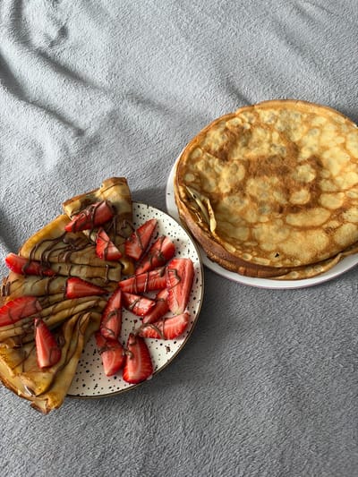 The image shows two plates of food placed on a grey fuzzy surface. One plate holds a stack of several golden-brown pancakes. The second plate contains folded pancakes topped with sliced strawberries and drizzled with chocolate sauce. The location is Новинки, Russia.