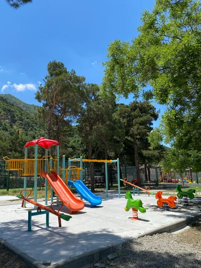 The image shows a playground in Mtskheta, Georgia, with a bright blue sky and green trees. The playground features several pieces of equipment, including orange and blue slides, a seesaw, and spring riders shaped like horses. In the background, a mountain covered in trees is visible. The ground is paved with concrete, and there are gravel areas around the playground. The scene is well-lit, suggesting it is daytime. There are no people visible in the image.