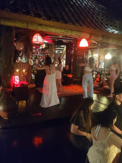 The image shows an indoor space, possibly a lounge or bar, located in Lokaserena, Indonesia, with a dark wooden floor and an open-air structure supported by wooden beams. The space is illuminated by red lanterns and other light sources, creating a warm and festive ambiance.
Several people are present in the scene. A woman in a long white dress is dancing with her arms raised. Other women are also dancing or standing nearby, some wearing casual attire like crop tops and shorts or light-colored pants. A man is seated, playing a hand drum, possibly providing musical accompaniment for the dancers. Other people are seated on sofas or cushions, observing the scene or engaged in conversation.
A large, decorative wooden mask with illuminated eyes sits on a platform in the background, adding a unique cultural touch to the setting. Other decorative elements, such as potted plants and hanging ornaments, contribute to the ambiance of the space. The overall scene suggests a lively social gathering or performance taking place in a bohemian-style venue in Lokaserena, Indonesia.