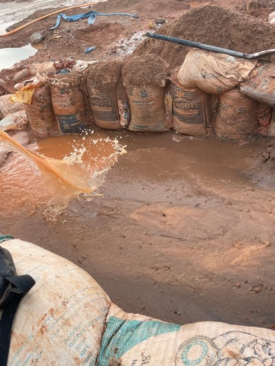 The image depicts a scene of construction materials, specifically bags of cement, situated in a muddy environment. The bags, some appearing wet and covered in mud, are stacked. One bag has visible text indicating "ANGOTE LIMESTONE CEMENT," and another states "SOKOTO CEMENT." The ground is saturated with water, creating puddles and muddy patches, suggesting recent rainfall or a wet construction site. A shovel is also visible, leaning against a pile of what appears to be sand or soil next to the cement bags. In the foreground, a splash of water indicates some kind of disturbance in the muddy water, perhaps from pouring or movement. Another large, white woven bag with some blue markings is partially visible on the right side. The overall scene suggests an active, albeit possibly challenging, construction or material handling operation in an outdoor setting. The lighting and muted colors suggest it might be overcast or late afternoon. The presence of mud and water strongly hints at a wet weather condition. While no people are directly visible, the presence of the shovel and stacked materials implies human activity. The text on the bags, such as "Rayfield, Nigeria" (though not directly visible in the cropped sections, implied by the prompt's location context), would further ground the location. The scene conveys a sense of industriousness in potentially unfavorable weather conditions.