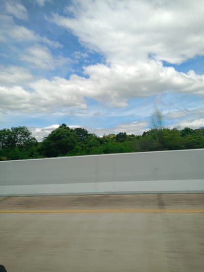 The image depicts a view from a moving vehicle on a highway near Seulimeum, Indonesia. The road is concrete with a solid yellow line marking the edge. A gray concrete barrier runs along the side of the road, separating it from a green tree line. The sky is blue with scattered white clouds. The motion blur indicates the vehicle is in motion.