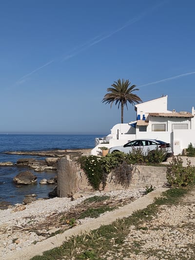 The image shows a coastal scene in Rotes, Spain. A white, modern house with a palm tree in front sits atop a low cliff above the sea. A grey car is parked in front of the house, partially obscured by bushes. The sea is calm and blue, with a rocky shoreline. A stone wall and natural vegetation, including flowering succulents, separate the house from a gravelly path. The sky is clear blue with two faint contrails.