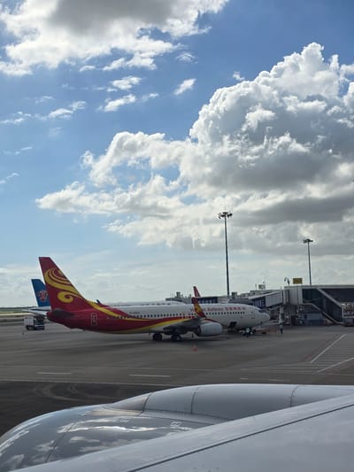 The image depicts an airport tarmac on a partly cloudy day. In the foreground, the metallic wing of an airplane is visible, reflecting the sky. Beyond this, a red and yellow airplane, identified by its tail fin and the text "Hainan Airlines" on its fuselage, is parked at a gate. Another airplane, with a blue tail fin, is parked behind it. Airport infrastructure, including jet bridges and lighting poles, is present. Several individuals in vests are seen on the tarmac, appearing to be airport personnel. The environment suggests a typical airport scene with planes and ground support operations underway. The location is specified as Bao'an District, China.